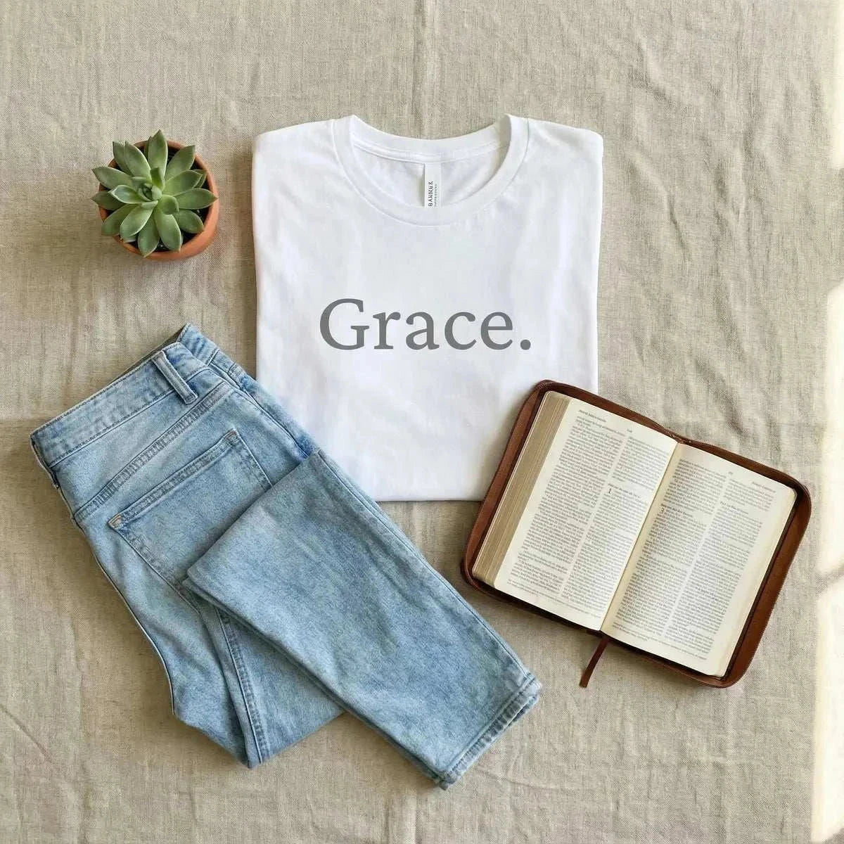 Flat lay of white Grace. Tee shirt on linen background surrounded by denim pants, a bible, and a succulent. 