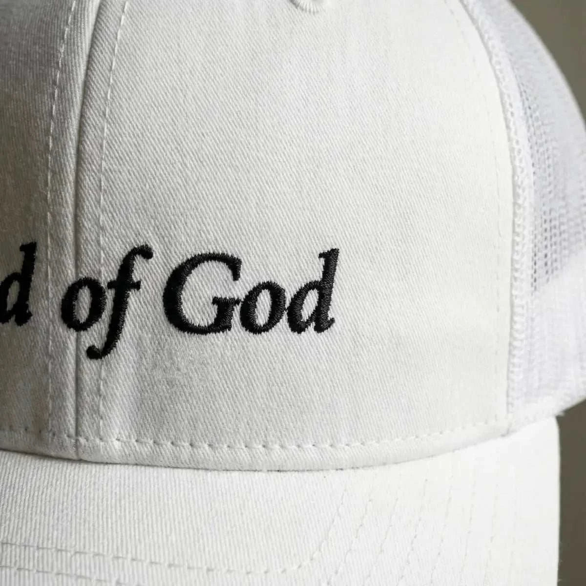 White cap with 'Child of God Trucker Hat' embroidered in black on a blurred background showing detail of texture and embroidery