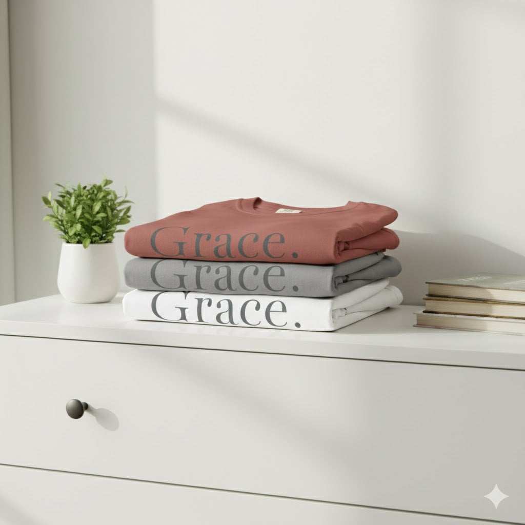 Stack of folded shirts with 'Grace. Tee' text on a white surface, next to a plant and books.