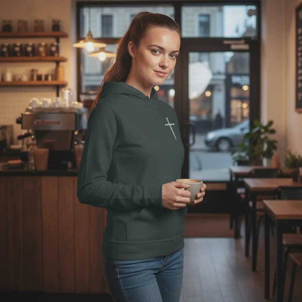 A woman wearing a Forest green hoodie with a small white cross logo on the chest, holding a coffee cup in a cafe