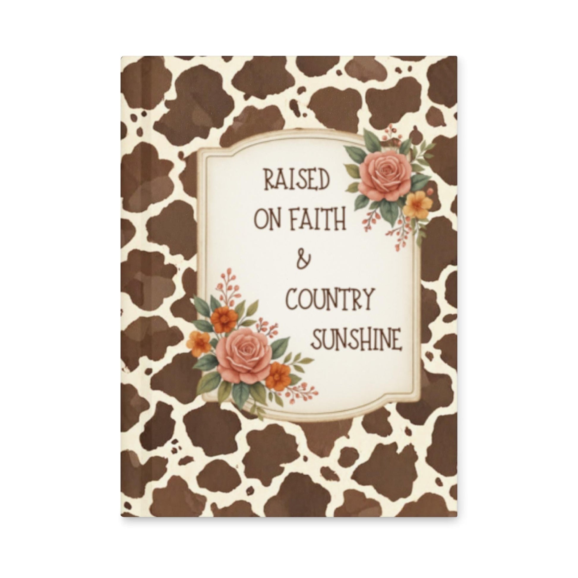 Front cover of a hardcover journal featuring a brown and white cow print pattern. A vintage cream-colored frame in the center reads "RAISED ON FAITH & COUNTRY SUNSHINE" in rustic brown text. The frame is decorated with peach and orange roses with green leaves.