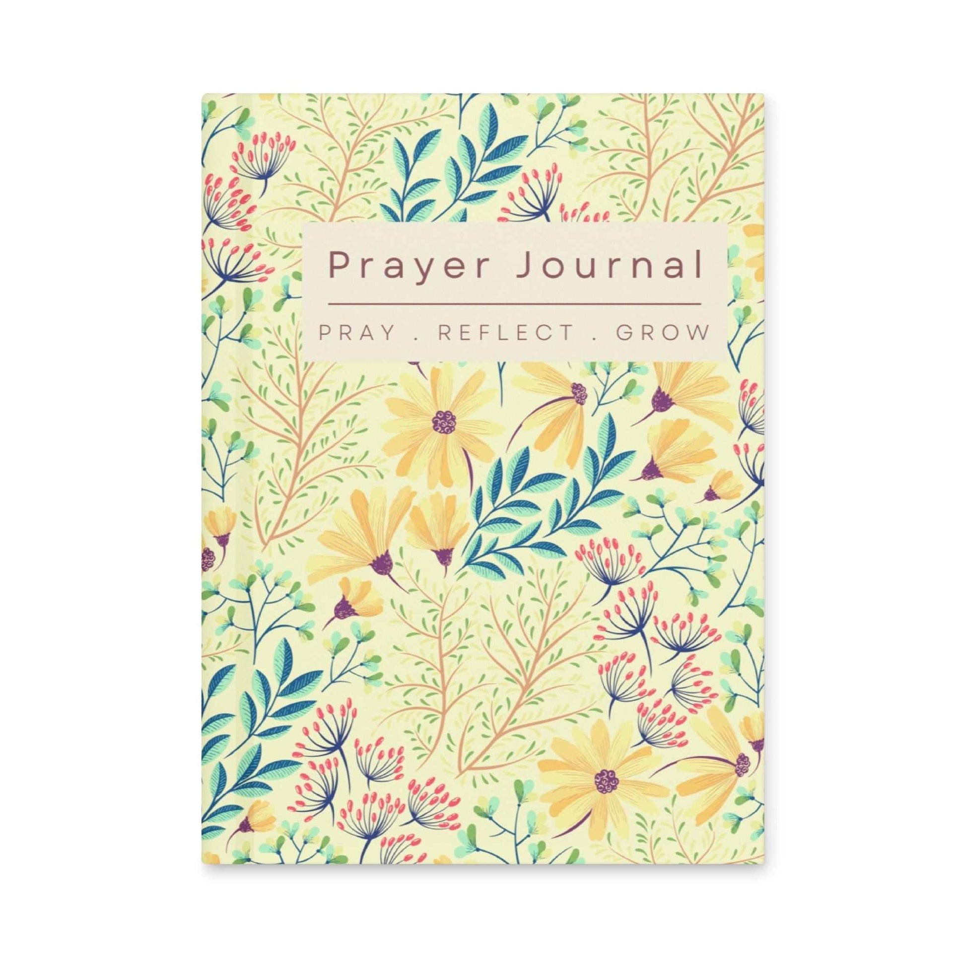 Front cover of floral hardcover journal featuring a vibrant pattern of yellow daisies, blue leaves, and red berries on a light yellow background. A beige rectangle in the center contains the words PRAYER JOURNAL, PRAY. REFLECT. GROW in capital letters.