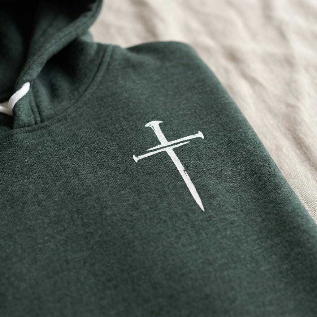 Close-up detail of a forest green hoodie showing a small white cross graphic printed on the chest, with soft fabric texture visible.