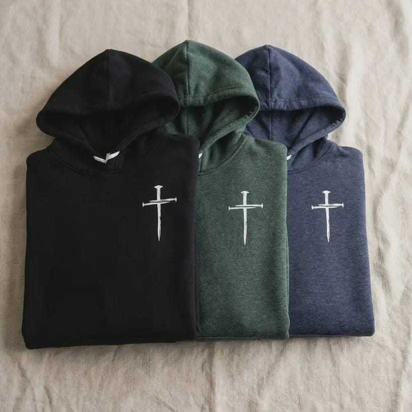 Three folded hooded sweatshirts are arranged side-by-side on a light beige textured fabric surface. From left to right, the hoodies are black, dark green, and dark blue. Each sweatshirt features a small, white, stylized cross graphic on the left side of the chest.
