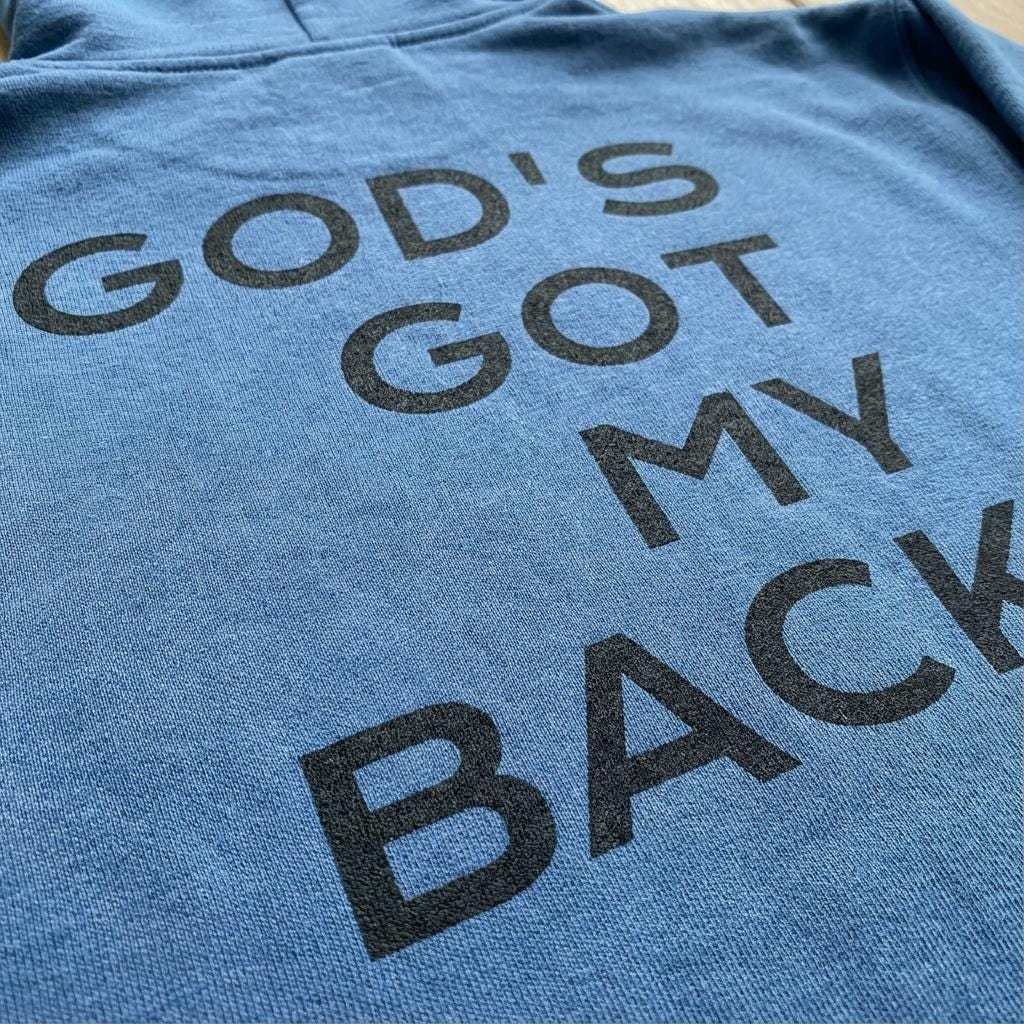 Close-up view of textured blue fabric, likely from a sweatshirt, featuring the phrase "GOD'S GOT MY BACK" printed in black, textured letters.