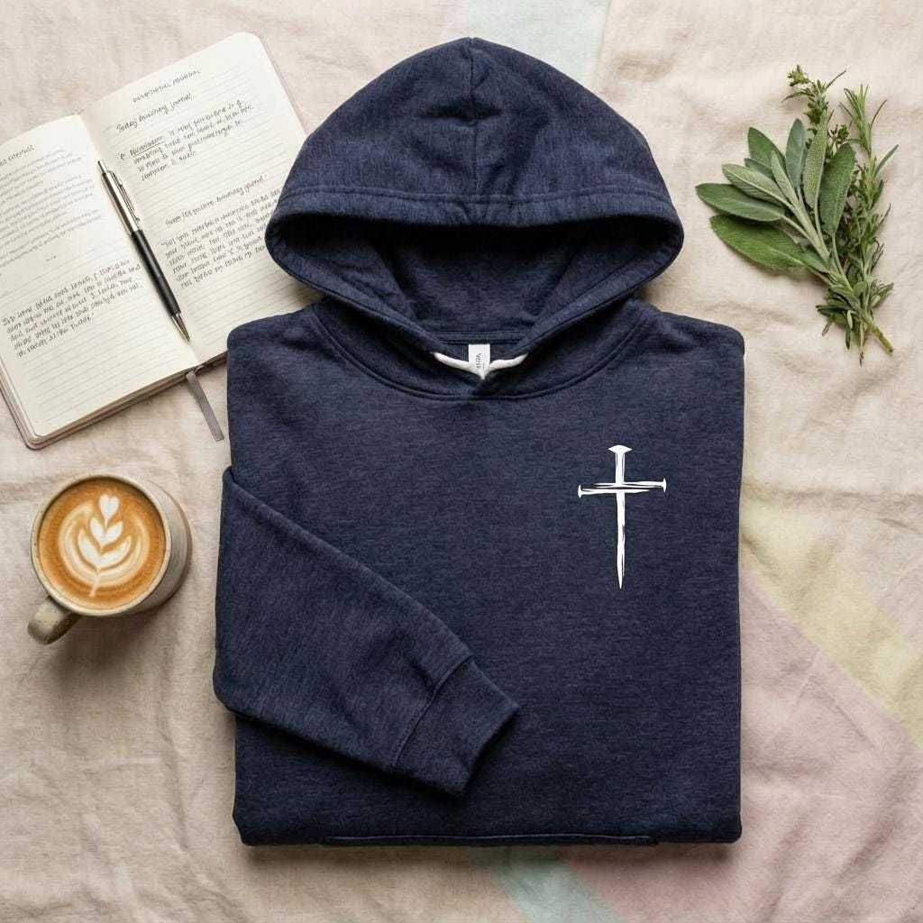 A top-down photograph of a folded, navy blue hooded sweatshirt with a small, white cross graphic printed on the right side of the chest area. The hoodie is neatly arranged on a textured fabric surface. To the left of the sweatshirt, there is an open spiral-bound journal with handwritten text and a silver pen resting on its pages. Below the journal is a ceramic mug filled with a latte, featuring a heart latte art design on the foam. To the right of the hoodie, a small bundle of fresh green herbs