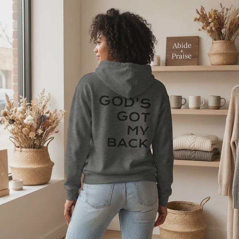 A woman with curly dark hair is standing and facing away from the camera, wearing a dark gray hooded sweatshirt. The back of the hoodie features the phrase "GOD'S GOT MY BACK" printed in large, bold, black capital letters. She is in a brightly lit room with a wooden shelf displaying dried plants, mugs, and a sign that reads "Abide + Praise".